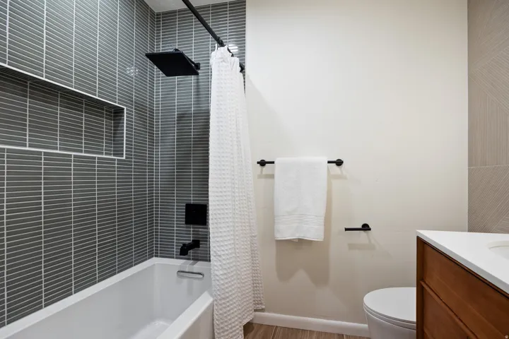 Bathroom featuring a white tub with a matte black rain shower head and fixtures, grey horizontal tile surround with a recessed niche, wood-finish flooring, and a vanity with a white countertop