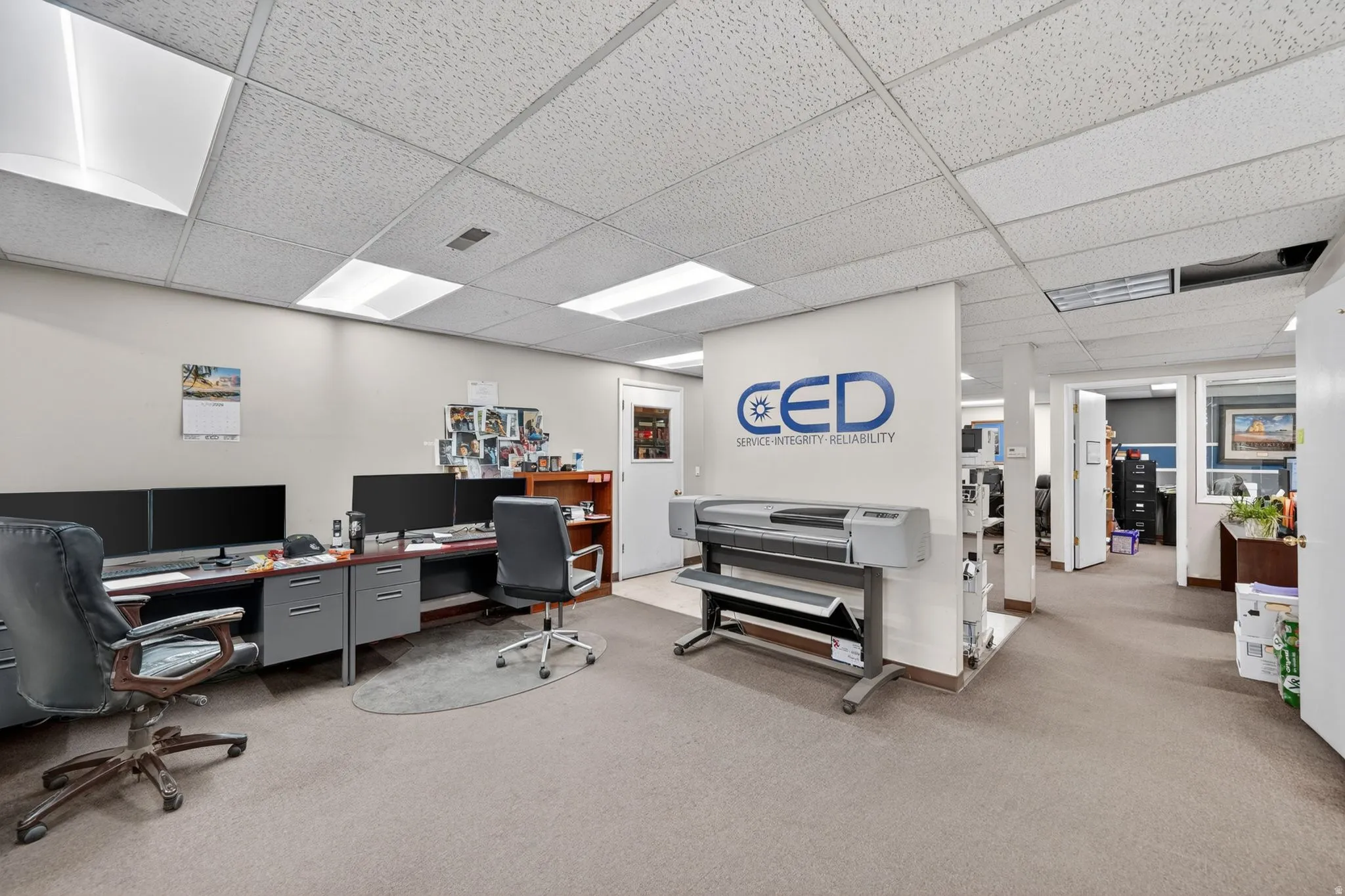 Open-plan office space featuring carpet flooring and a suspended acoustic ceiling with recessed lighting