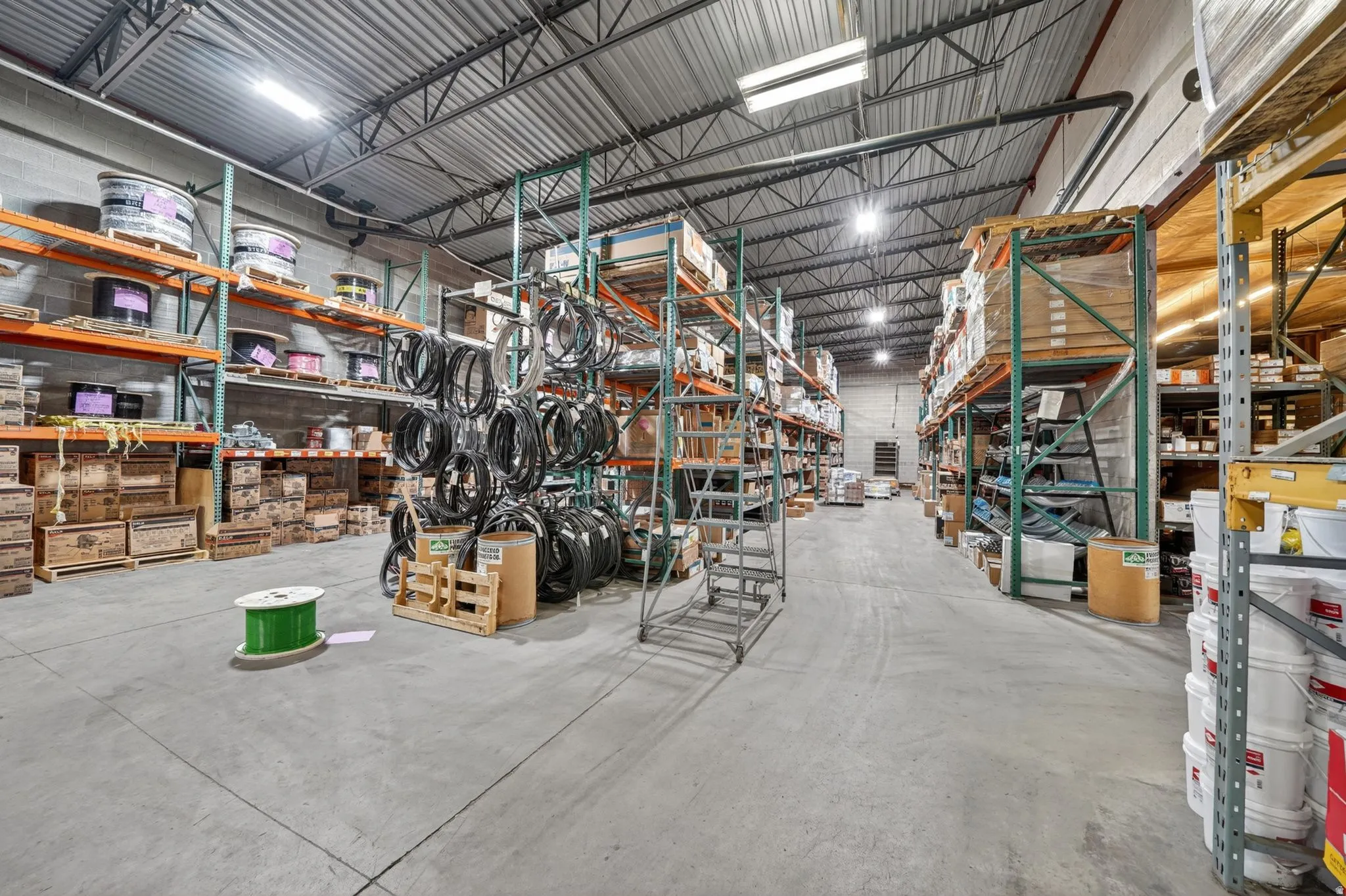 Spacious warehouse interior featuring high ceilings with exposed metal trusses, concrete flooring, and extensive metal shelving systems