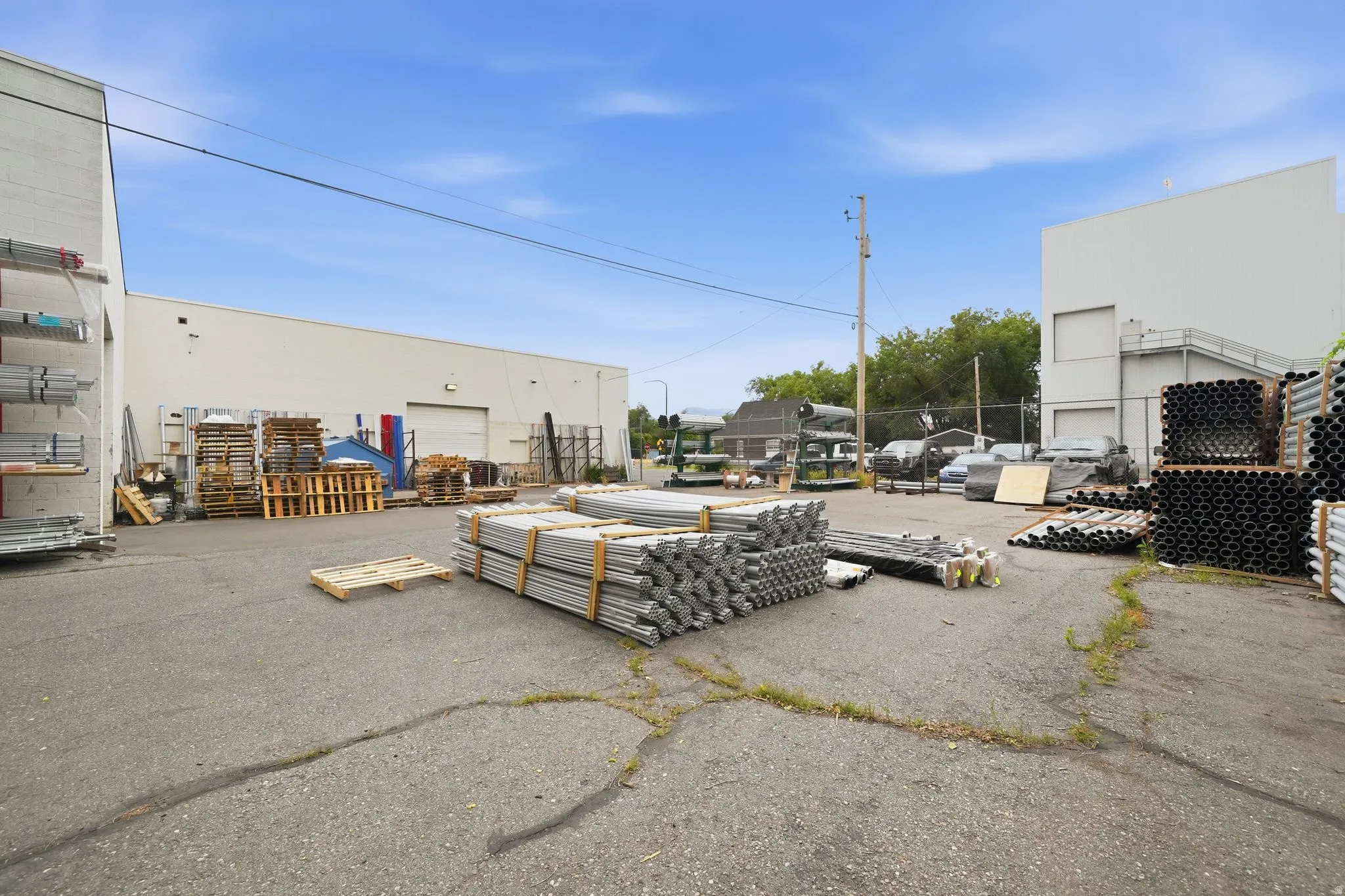 Paved outdoor storage area featuring two large light-colored industrial buildings, multiple overhead doors, and exterior wall-mounted shelving