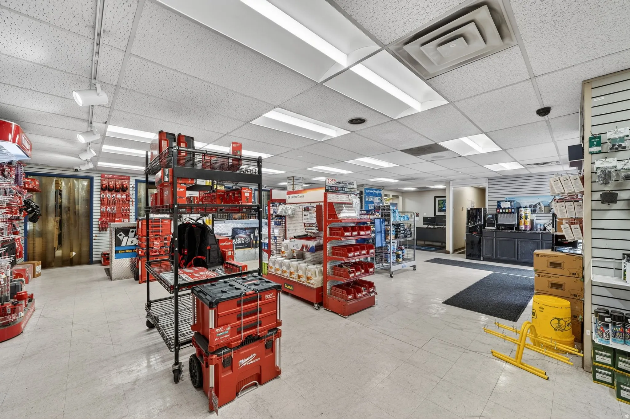 Retail interior featuring white floor tiles, a suspended ceiling with integrated fluorescent lighting, track lighting fixtures, and slatwall paneling