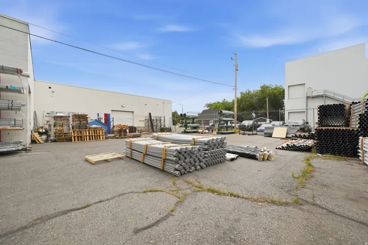 Paved outdoor storage area featuring two large light-colored industrial buildings, multiple overhead doors, and exterior wall-mounted shelving