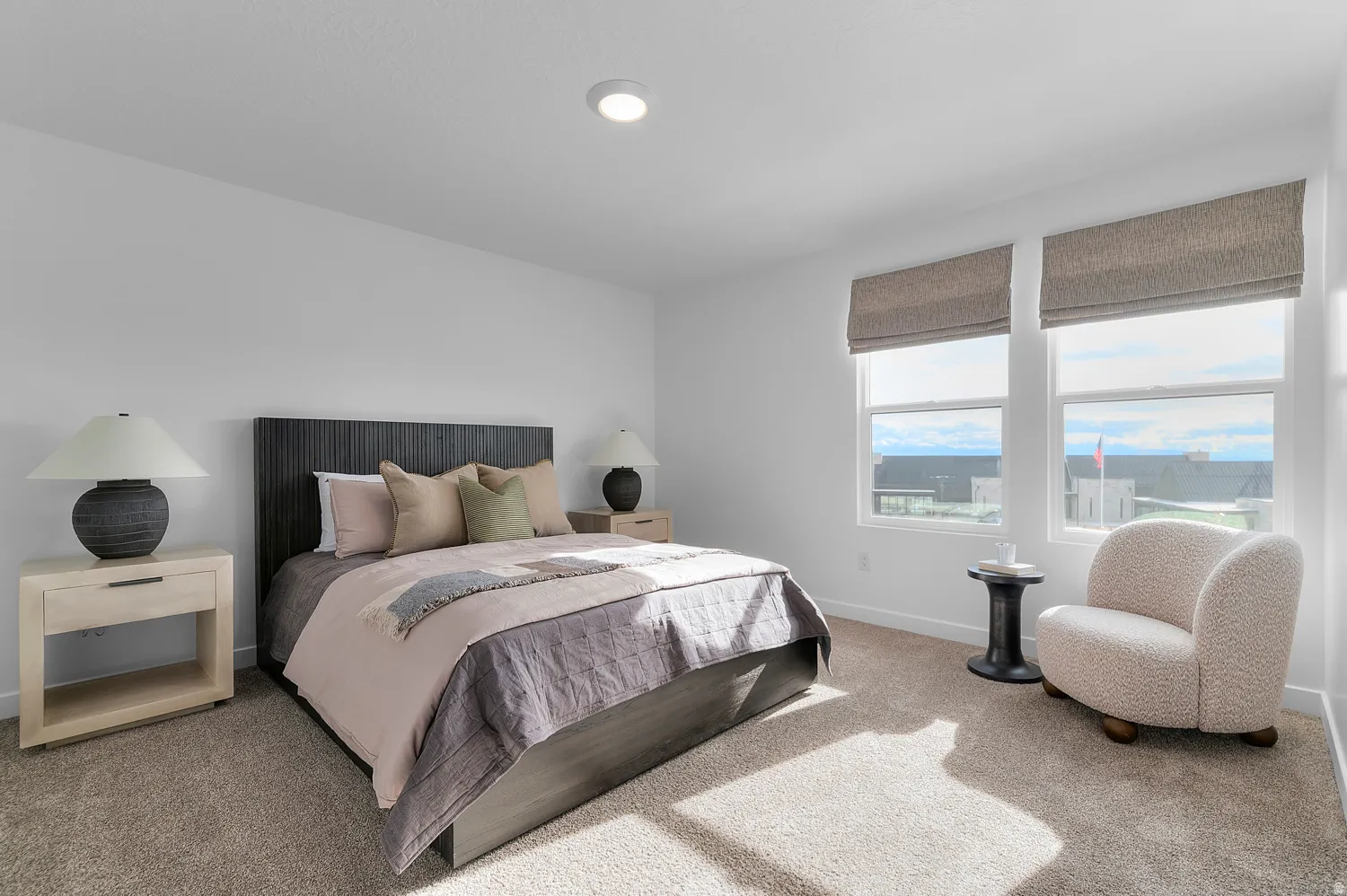 Carpeted bedroom featuring a textured dark wood headboard, two large windows with neutral-toned Roman shades, and recessed ceiling lighting