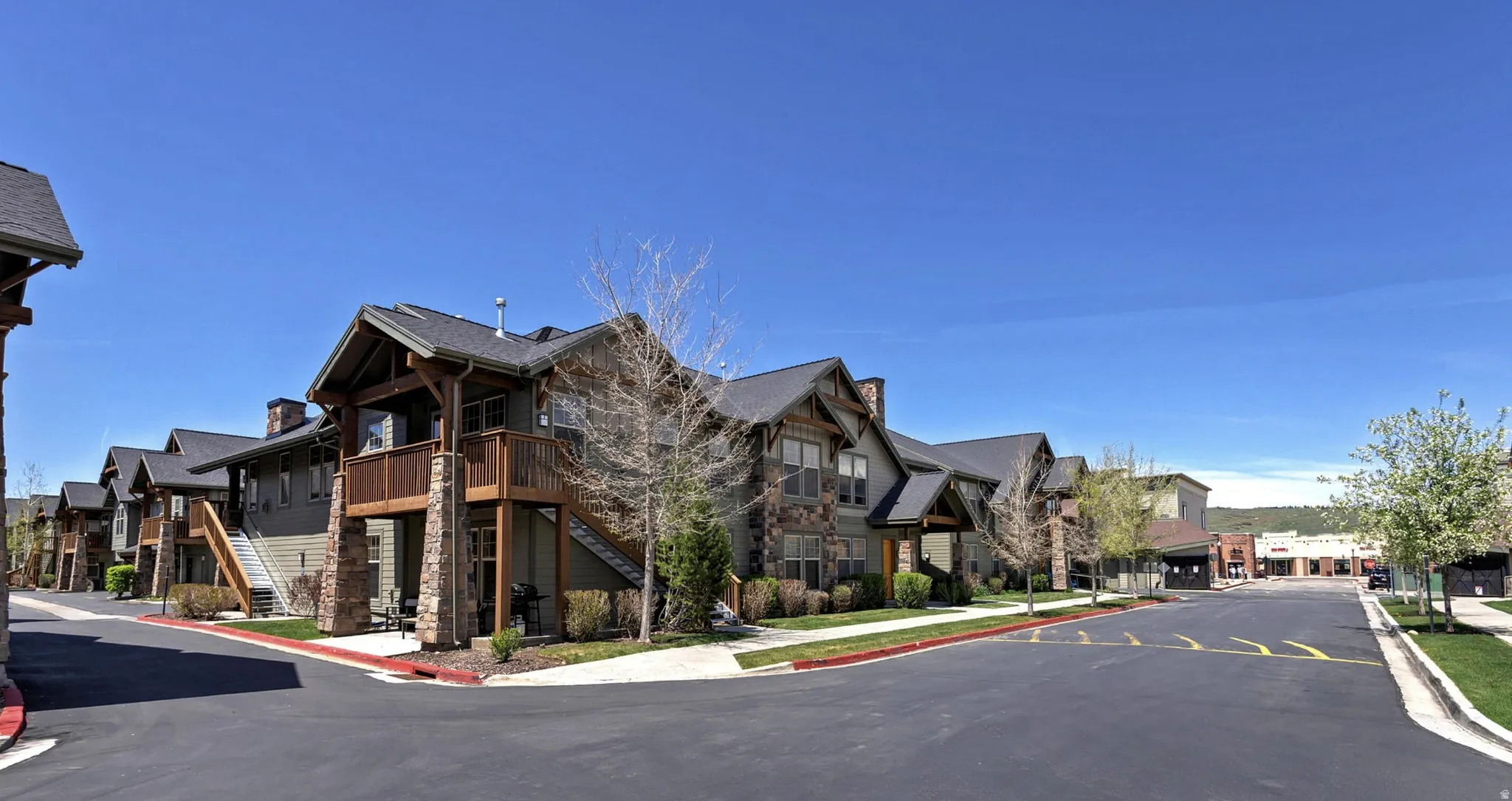 Multi-story residential buildings featuring stone and siding exteriors, dark shingle roofs, wood accents on balconies and structural supports, and paved roadways with curbing