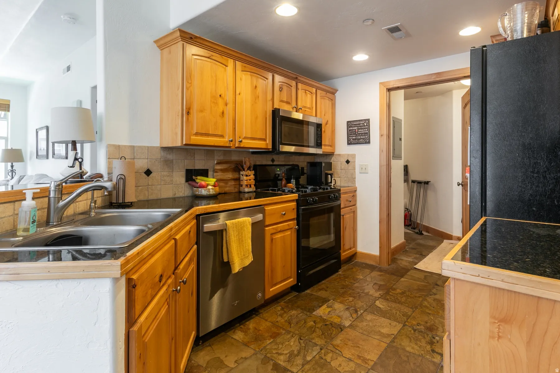 Kitchen featuring natural wood cabinetry, a stainless steel dishwasher, black appliances, a tile backsplash, and dark countertops