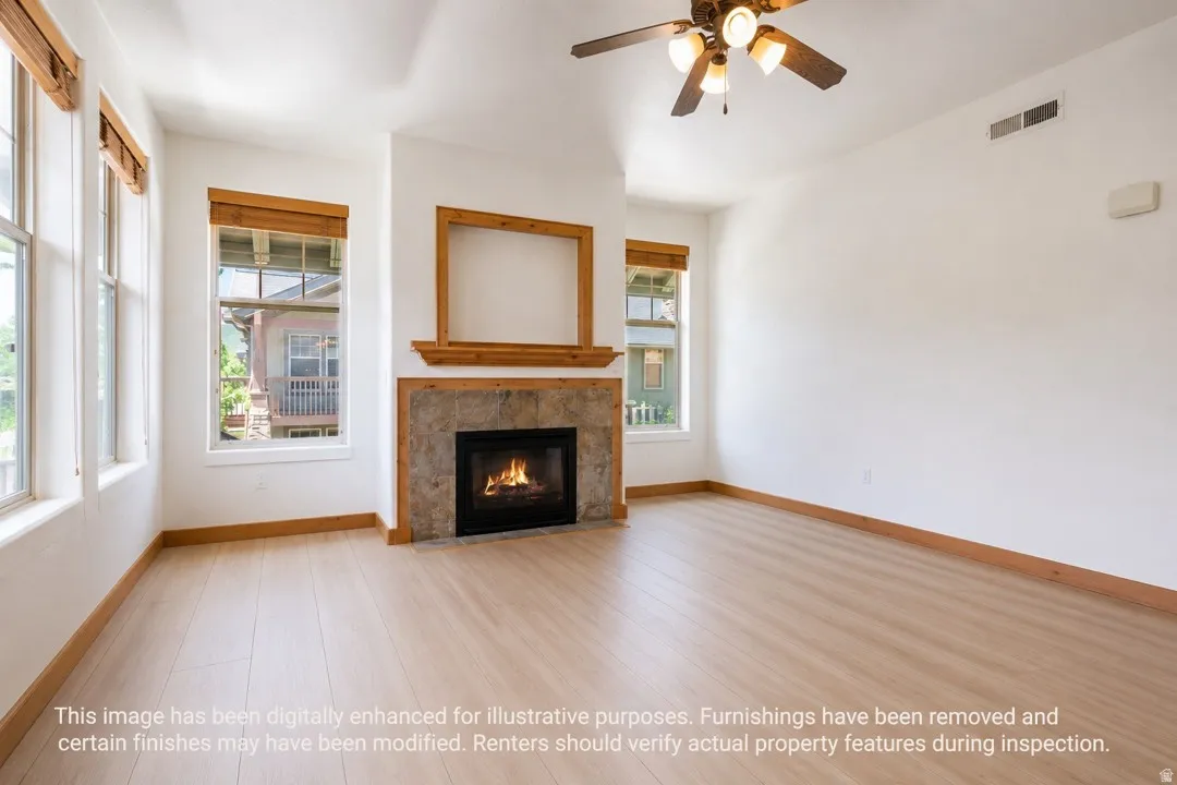 Spacious living area featuring light wood-finish flooring, a stone-faced fireplace with a wood mantle, and a ceiling fan with integrated lighting