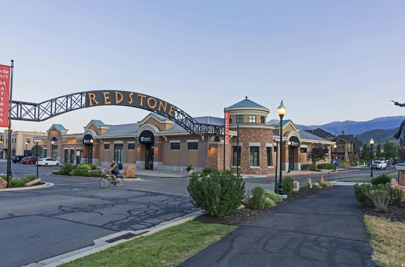 Commercial retail complex featuring a prominent arched "REDSTONE" entrance sign, a circular building with a domed roof and stone facade, multiple storefronts with awnings, pedestrian pathways, and integrated landscaping