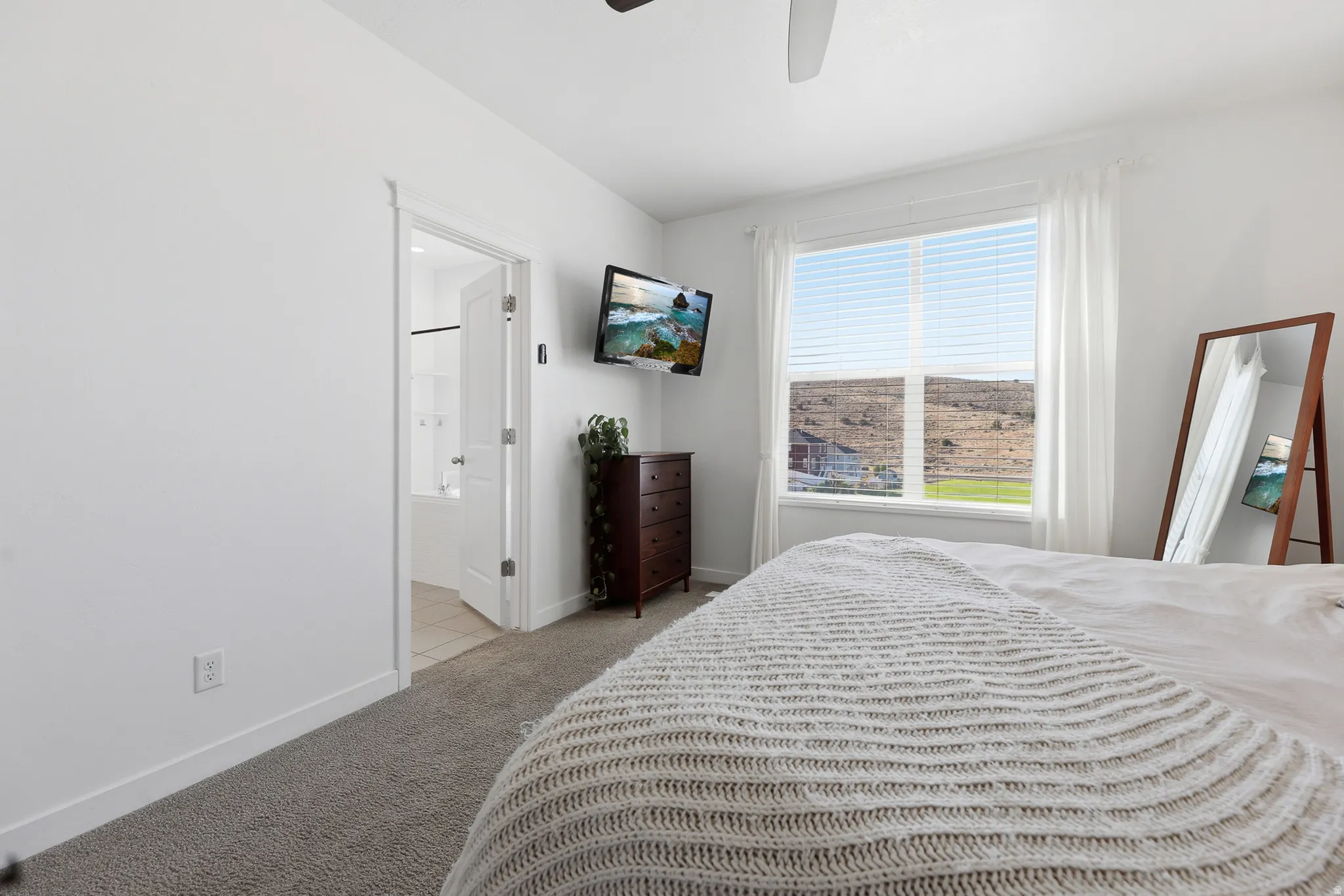 Carpeted room with white walls, a large window with horizontal blinds, and an open doorway revealing a bathroom with tile flooring and a bathtub