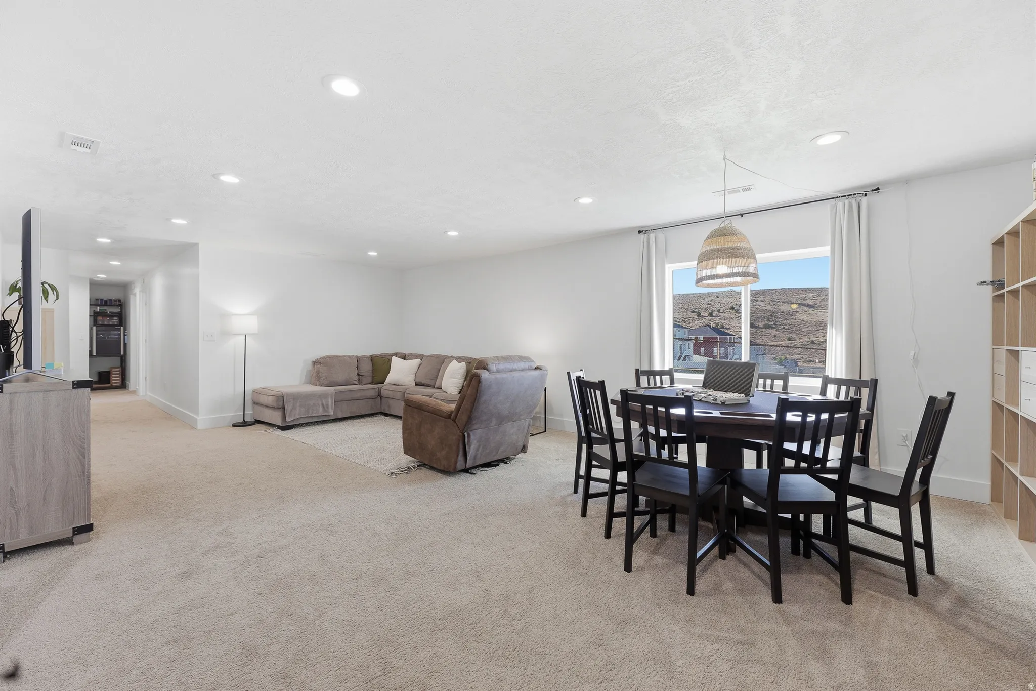 Spacious open-concept room featuring neutral-toned carpeting, ample recessed lighting, a large window with a distant landscape, and a built-in shelving unit