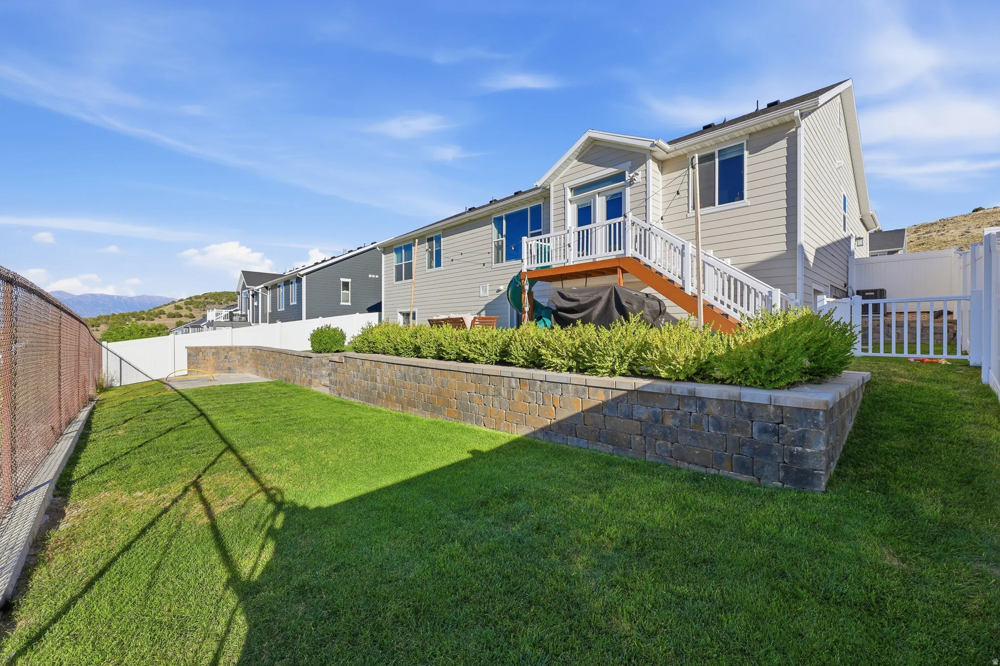 Rear exterior featuring a raised deck with white railings, a lower-level grass area, and a stone retaining wall with mature shrubbery