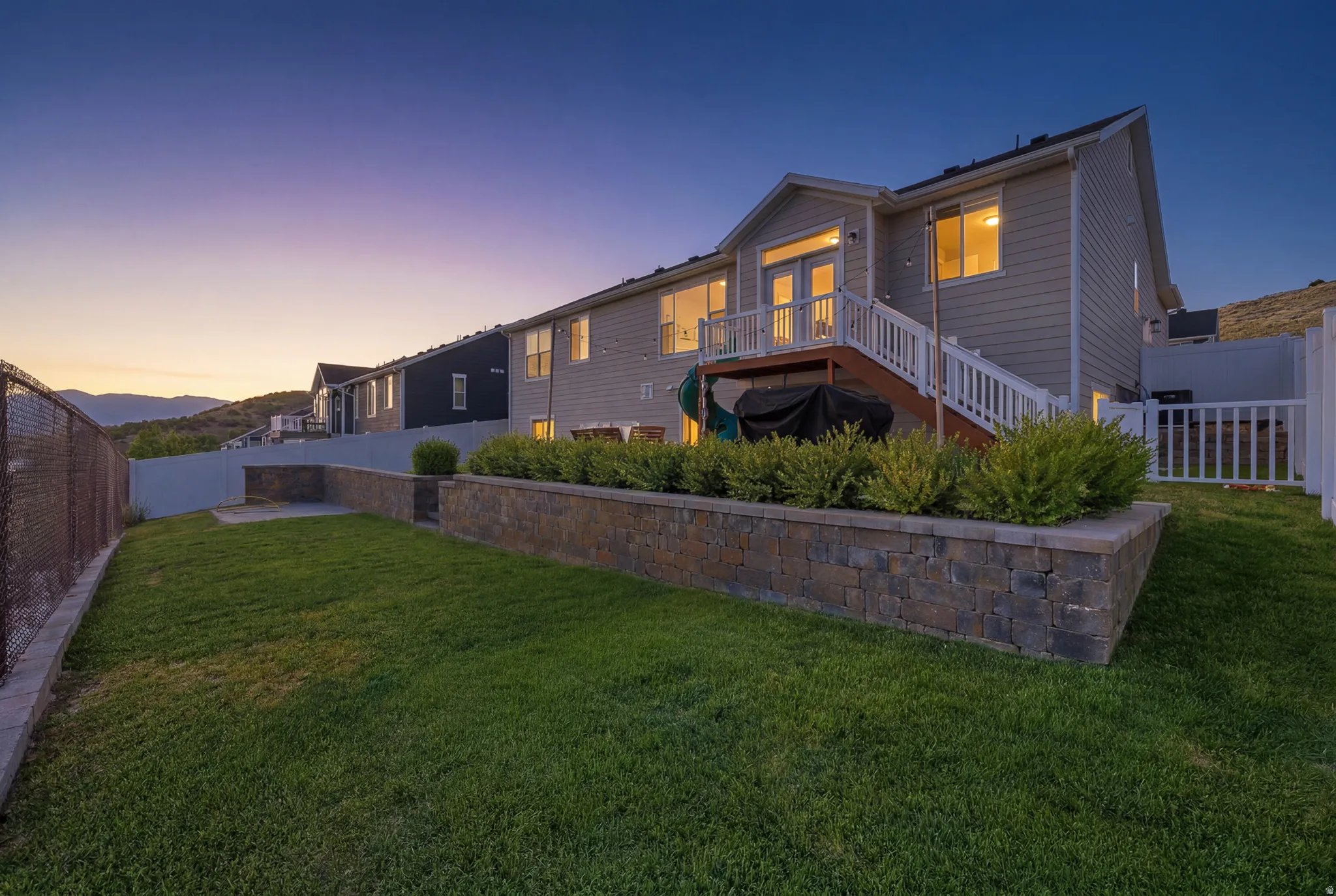 Expansive backyard featuring a manicured lawn, a multi-level retaining wall with integrated shrubbery, a chain-link fence, and a white picket fence