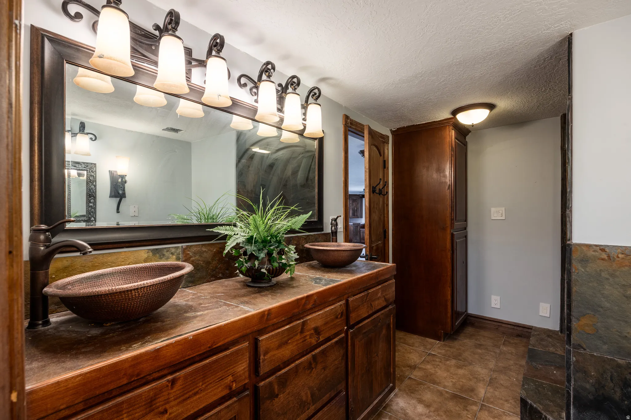 Dual vessel sinks with bronze fixtures, extensive vanity with wood-finish cabinetry, broad framed mirror, tiled flooring, and built-in storage