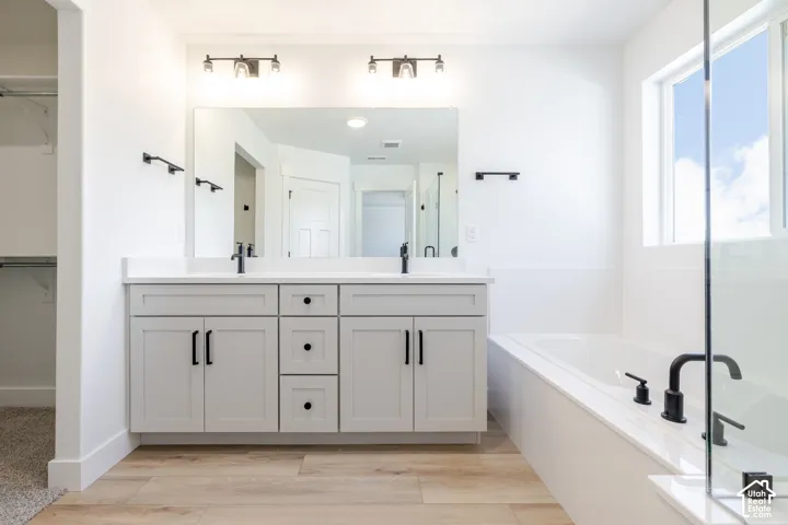 Dual-sink vanity with light-tone shaker cabinetry, quartz countertop, wood-finish flooring, drop-in soaking tub, and matte black fixtures