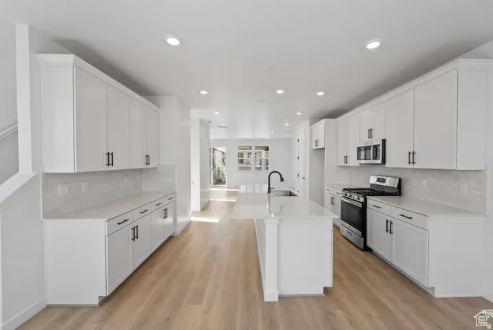 Spacious kitchen featuring white shaker cabinetry, light wood-finish flooring, a central island with an undermount sink, stainless steel appliances, and recessed lighting