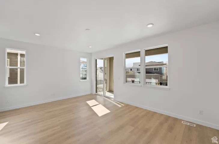 Spacious room featuring light wood-finish flooring, recessed lighting, multiple windows with white trim, and a sliding glass door leading to an exterior balcony
