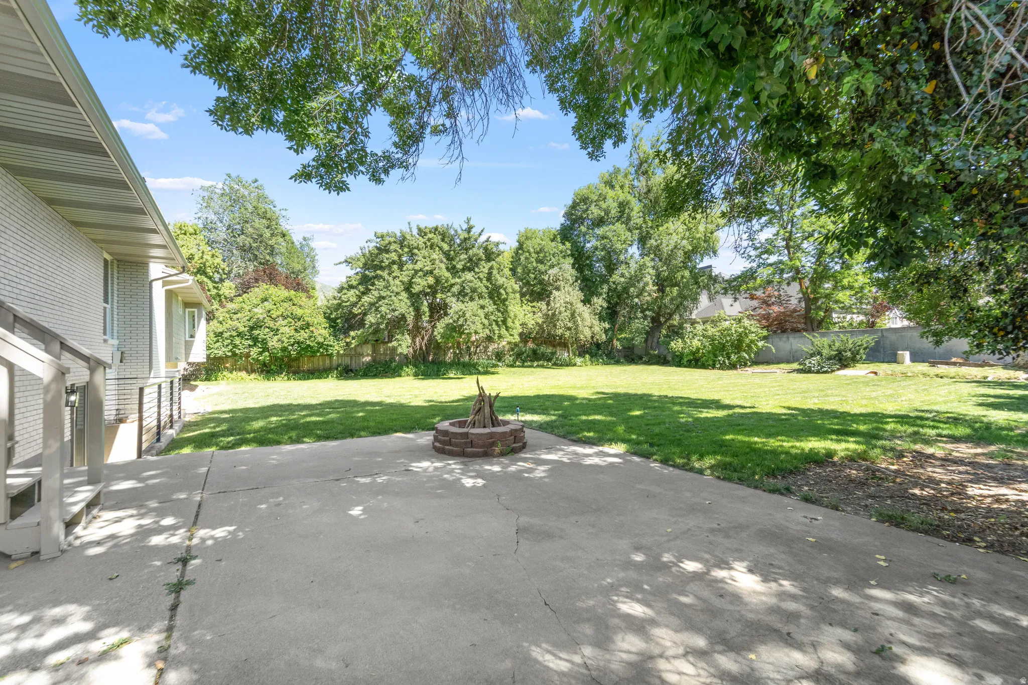 Expansive concrete patio offering a brick fire pit, a sprawling lawn, and mature perimeter trees