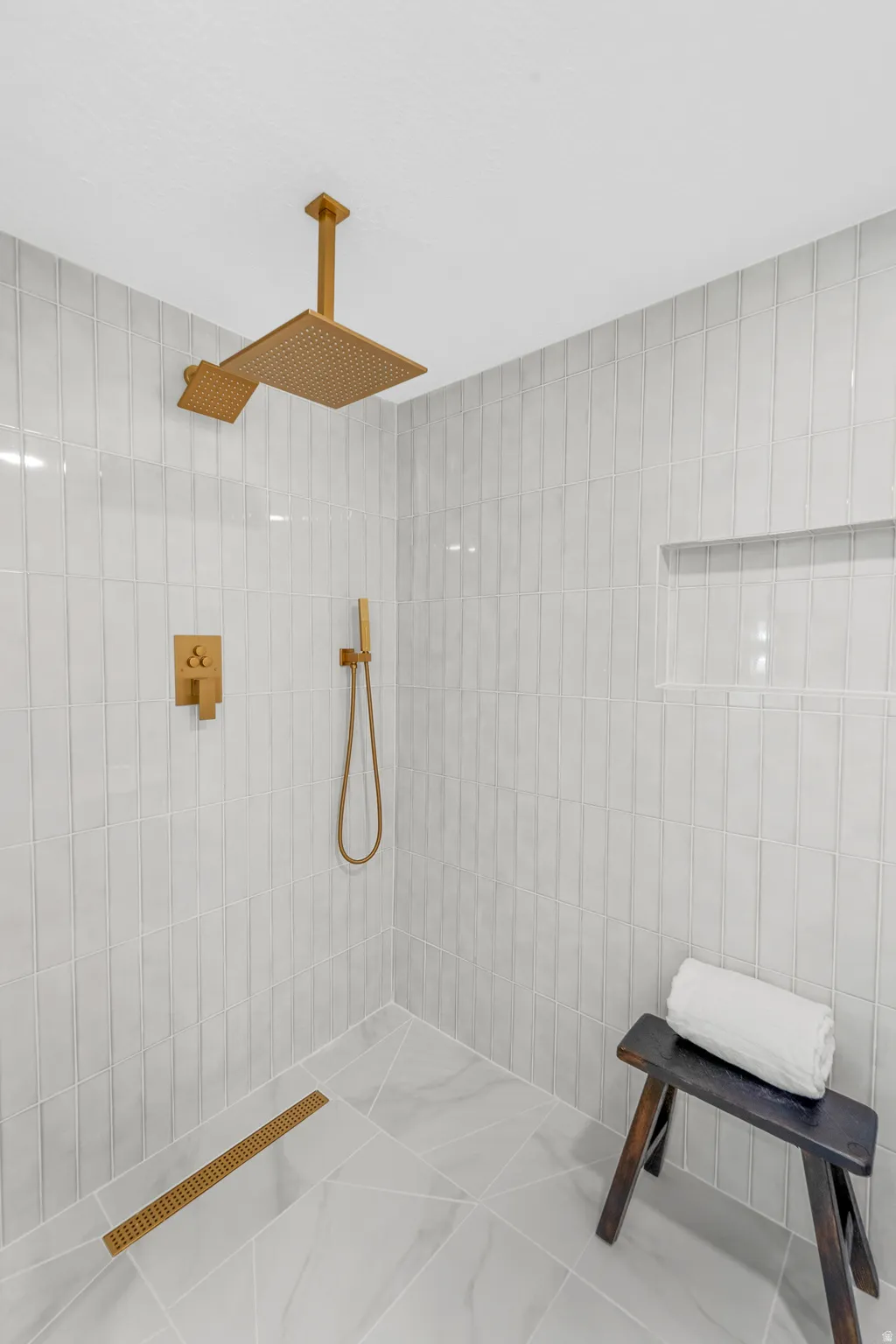 Walk-in shower featuring light-toned vertical tile walls, a ceiling-mounted gold-finish rainfall showerhead, a hand-held shower wand, and a linear shower drain