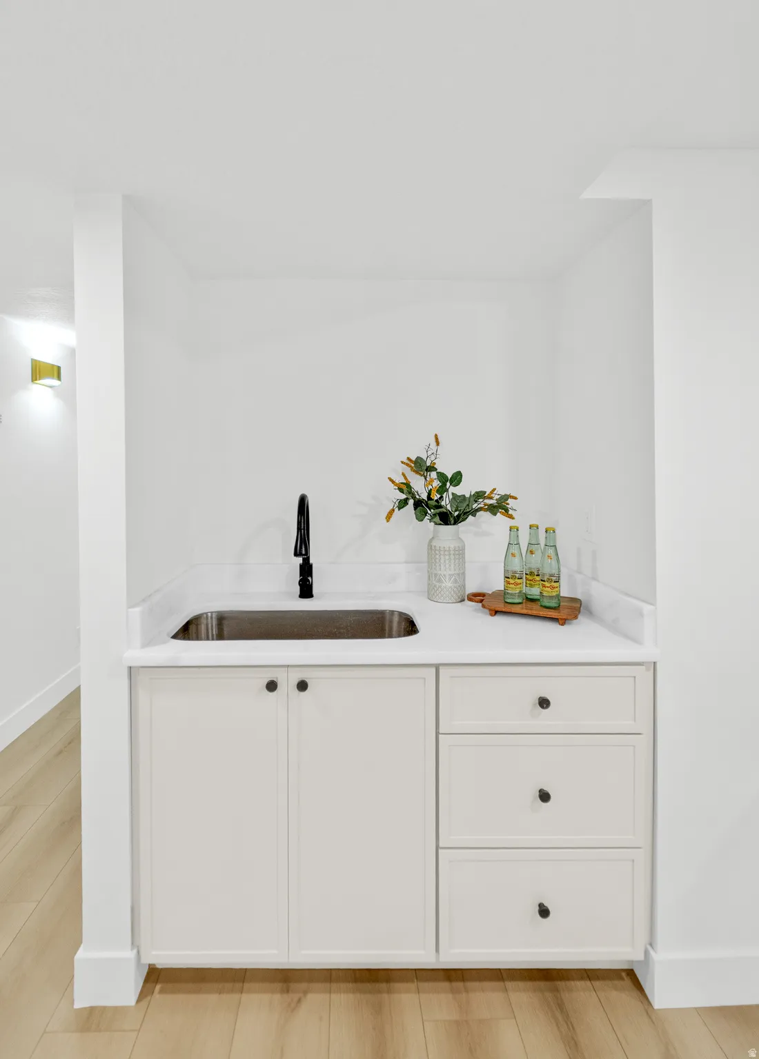 Built-in wet bar featuring a stainless steel undermount sink, black gooseneck faucet, light-toned cabinetry with dark hardware, and light-colored countertops with a subtle veining pattern