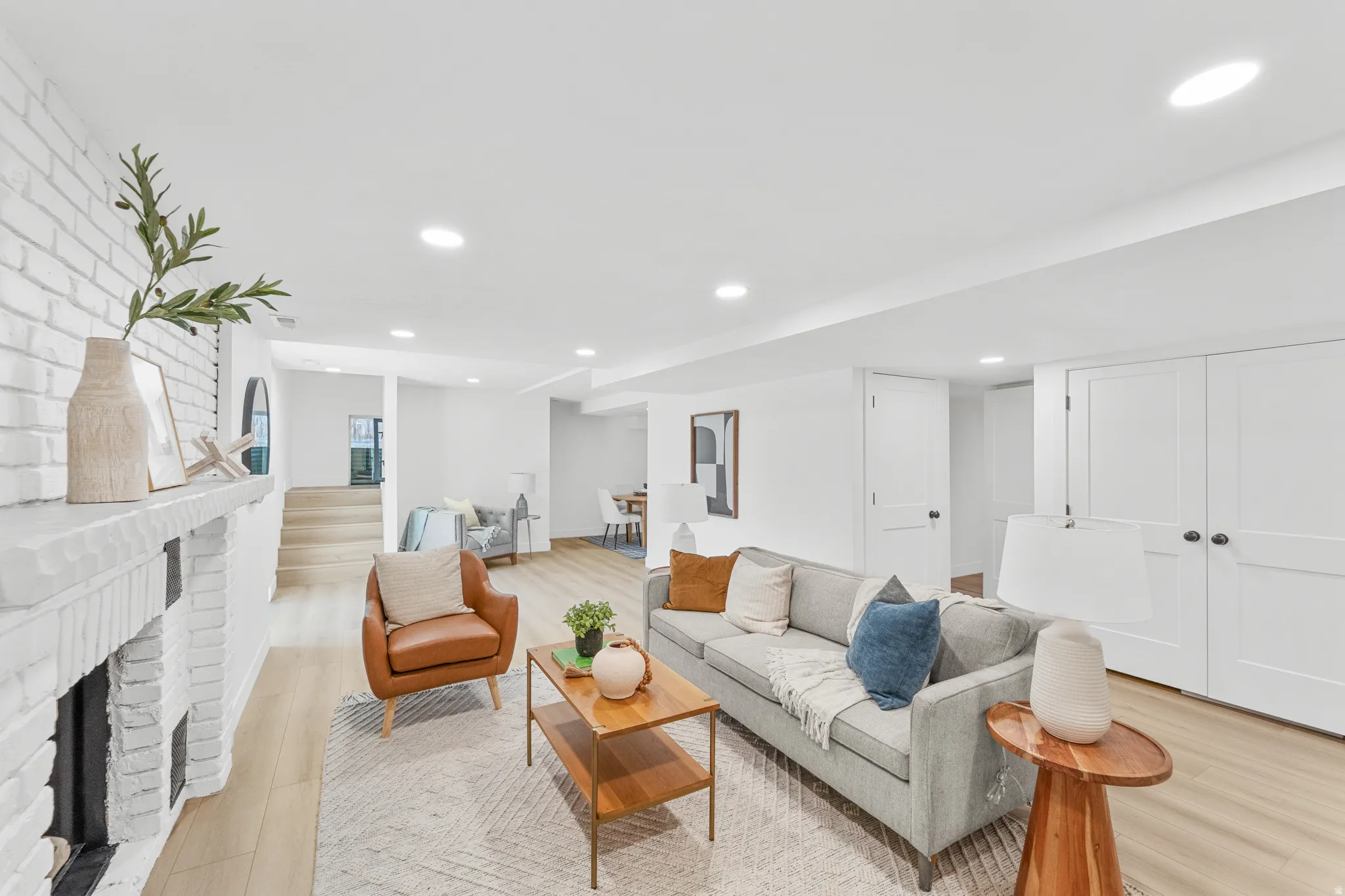 Open-concept living area featuring a whitewashed brick fireplace, wood-finish flooring, recessed lighting, white walls, and built-in closet doors