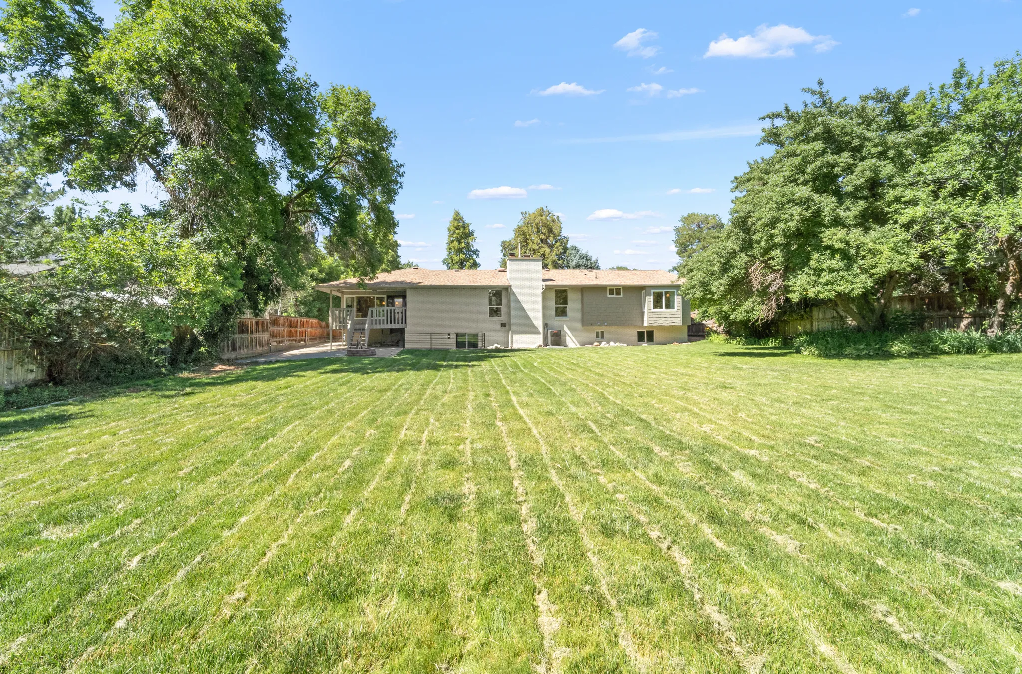 Expansive backyard featuring a large green lawn, mature perimeter trees, and a two-story residence with a covered deck and chimney
