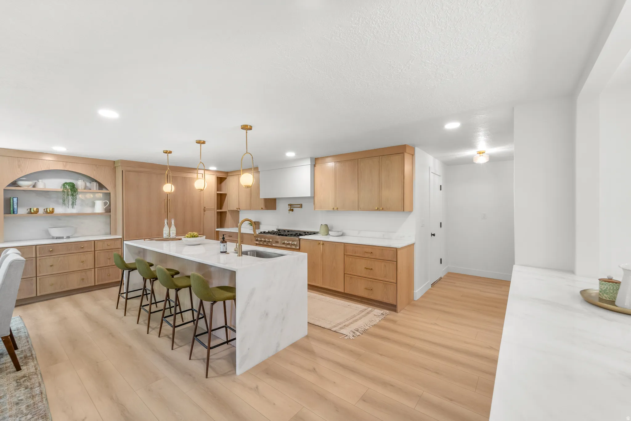 Spacious kitchen featuring a large waterfall island with integrated sink, light wood-finish cabinetry, built-in shelving, a professional-grade range, and recessed lighting throughout