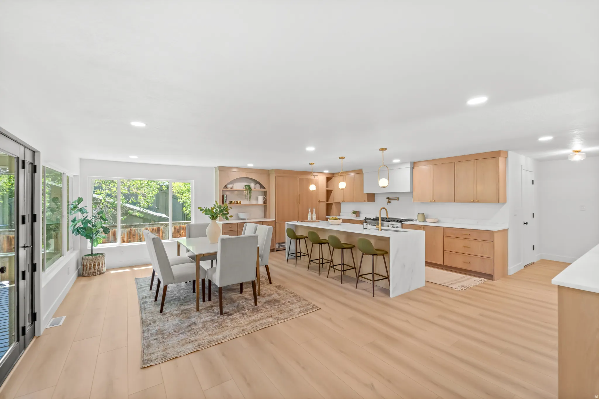 Open-concept living area featuring light wood-finish flooring, a modern kitchen with natural wood cabinetry and a waterfall island, recessed lighting, and expansive windows