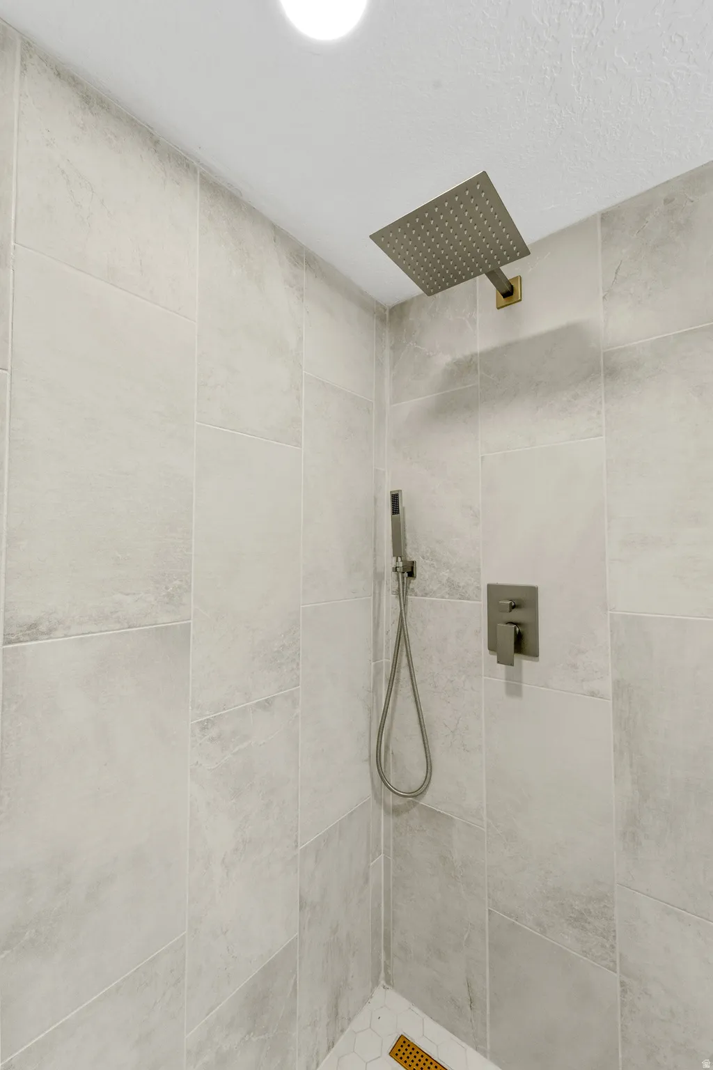 Spacious shower featuring light-toned large format wall tiling, a rain shower head, a handheld shower fixture, and a mosaic hex tile shower pan with a linear drain