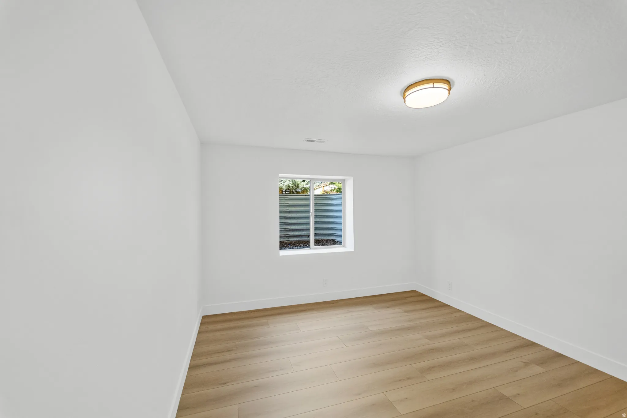 Room featuring light-toned wood-finish flooring, a contemporary ceiling light fixture, and a window with a corrugated window well