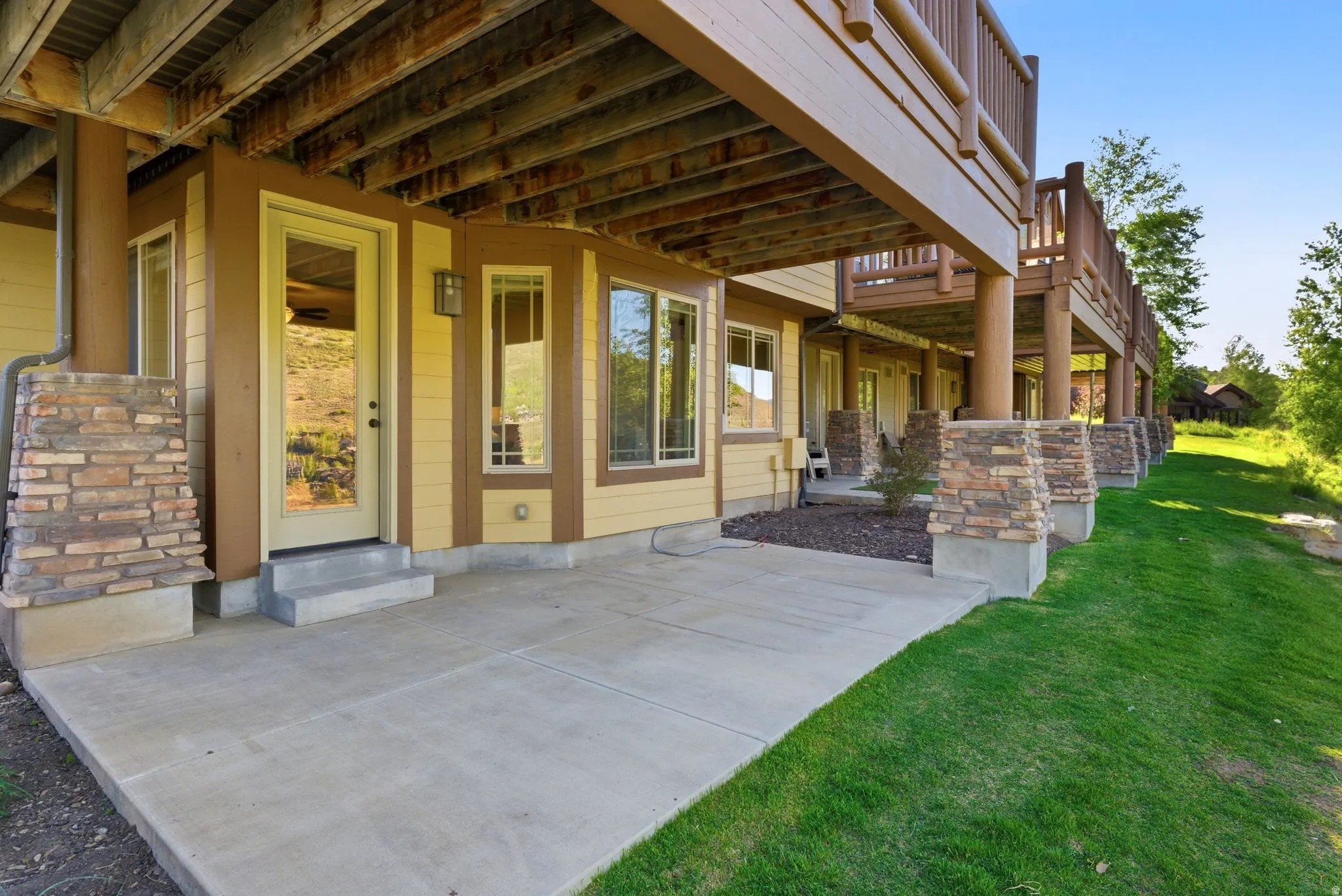 Spacious concrete patio beneath an upper-level deck, featuring a glass-paneled door, architectural windows, and stone-clad support pillars
