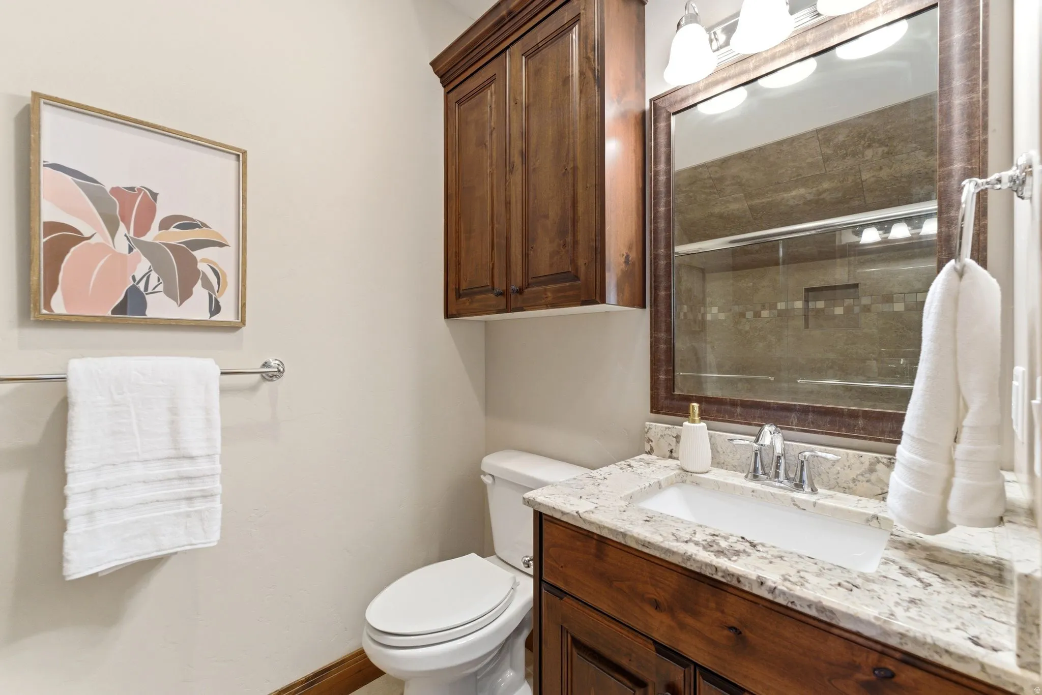 Bathroom featuring a white rectangular undermount sink set within a light-colored speckled countertop, wood-finish vanity cabinetry, a wood-framed mirror with an overhead three-light fixture, a toilet, and a wall-mounted wood-finish cabinet