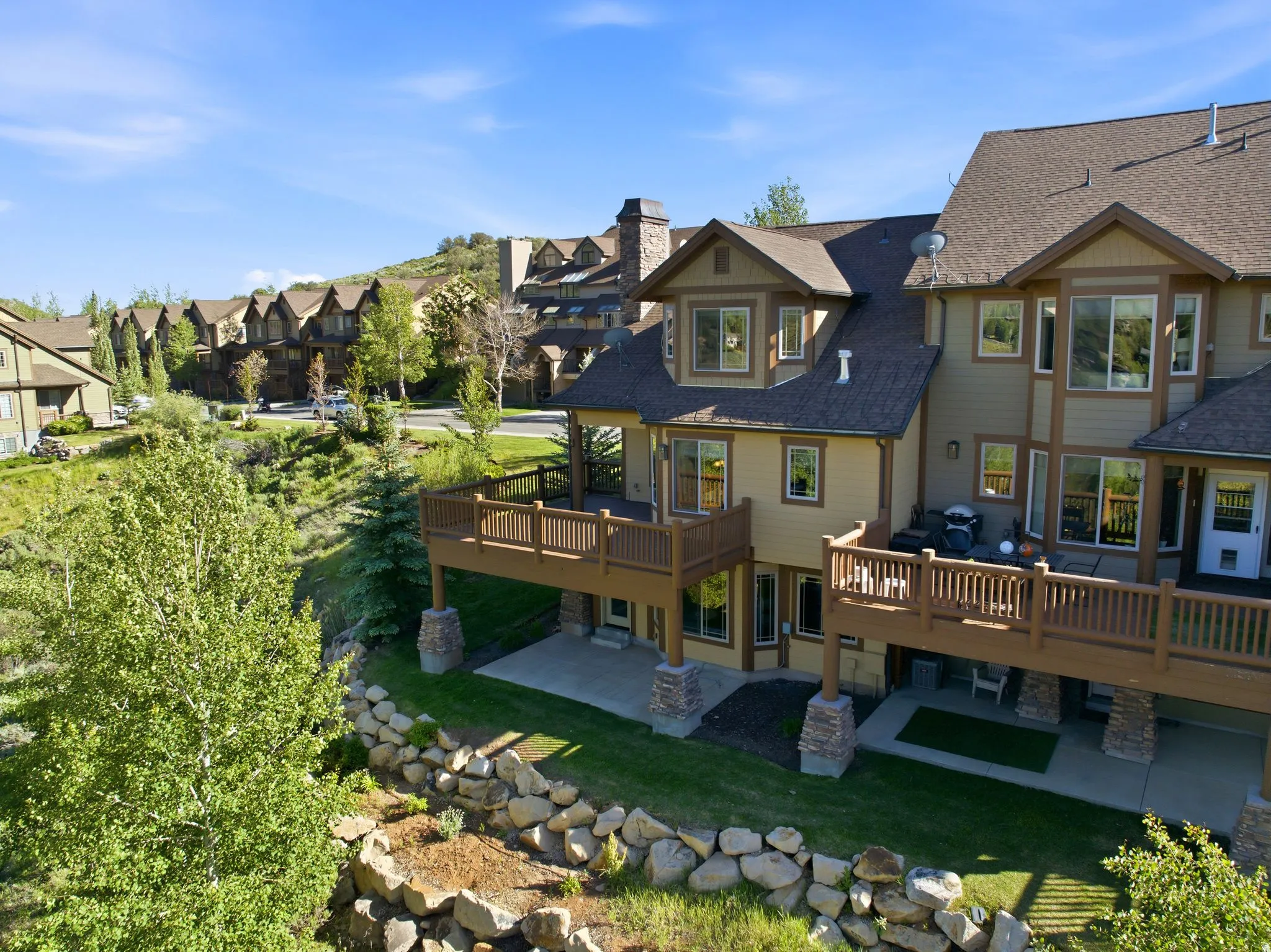 Exterior featuring multi-level decks with wood railings, a stone-clad chimney, an asphalt shingle roof, and a lower-level patio with concrete flooring