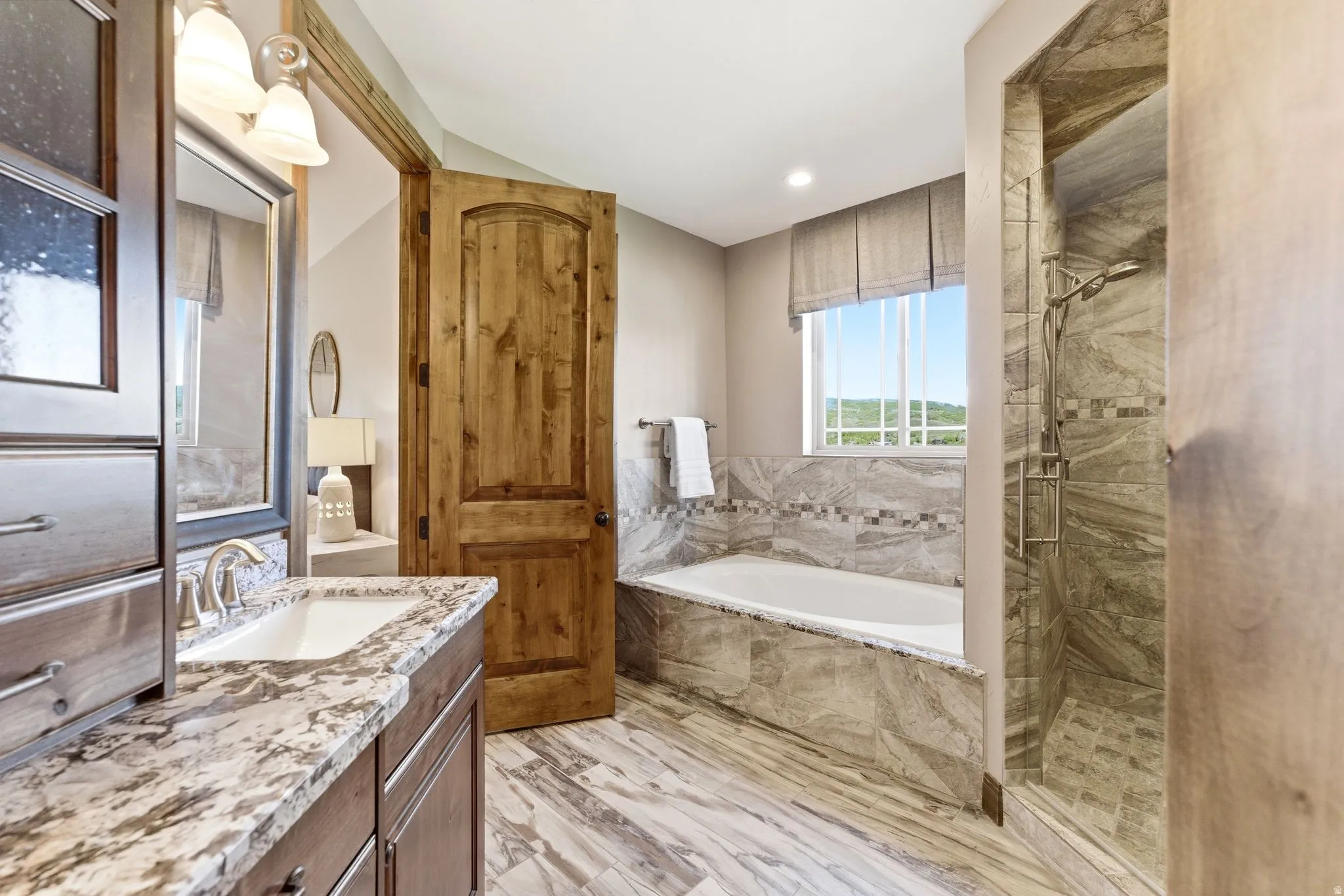 Bathroom featuring a built-in tub with tile surround, a separate tiled shower enclosure, a single vanity with stone countertop and integrated sink, wood-finish flooring, and a wood-panel door