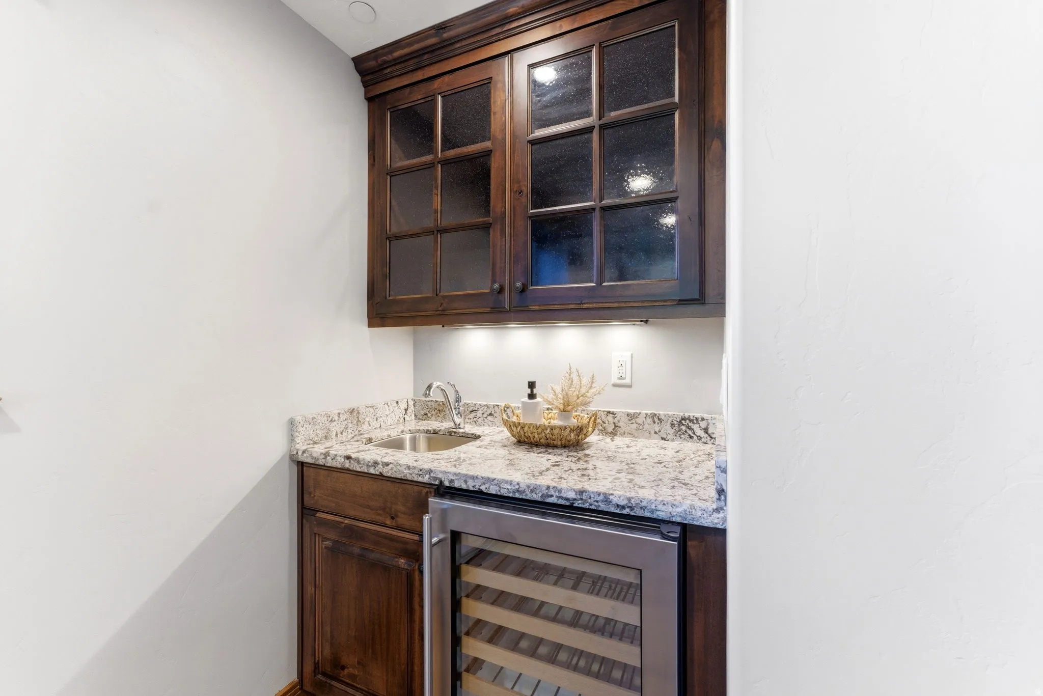 Wet bar featuring rich wood cabinetry, a stainless steel sink, granite countertop, glass-front upper cabinets, and an integrated beverage refrigerator