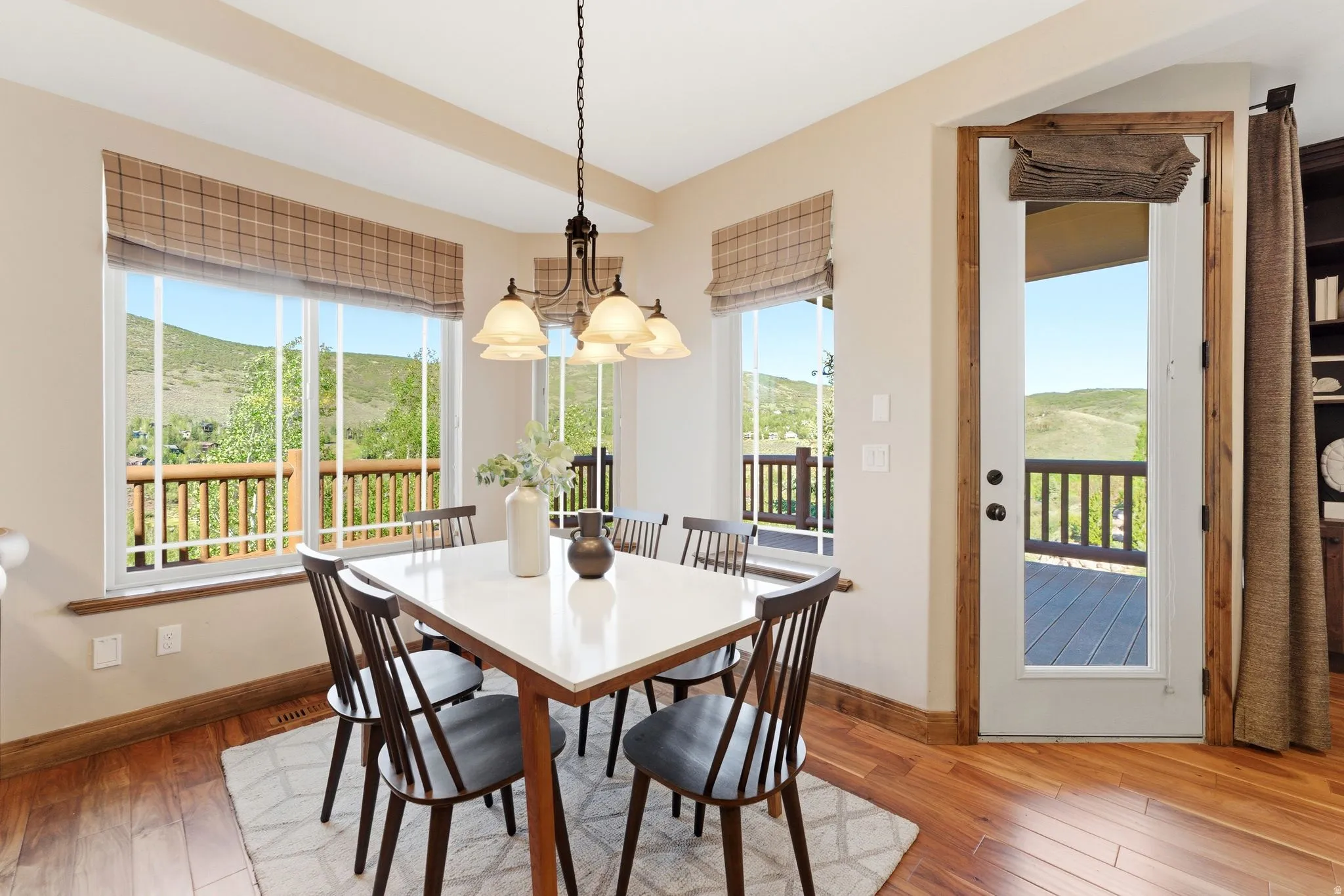 Bright dining area featuring hardwood flooring, multiple large windows with plaid roman shades, an overhead chandelier, and direct access to an exterior deck
