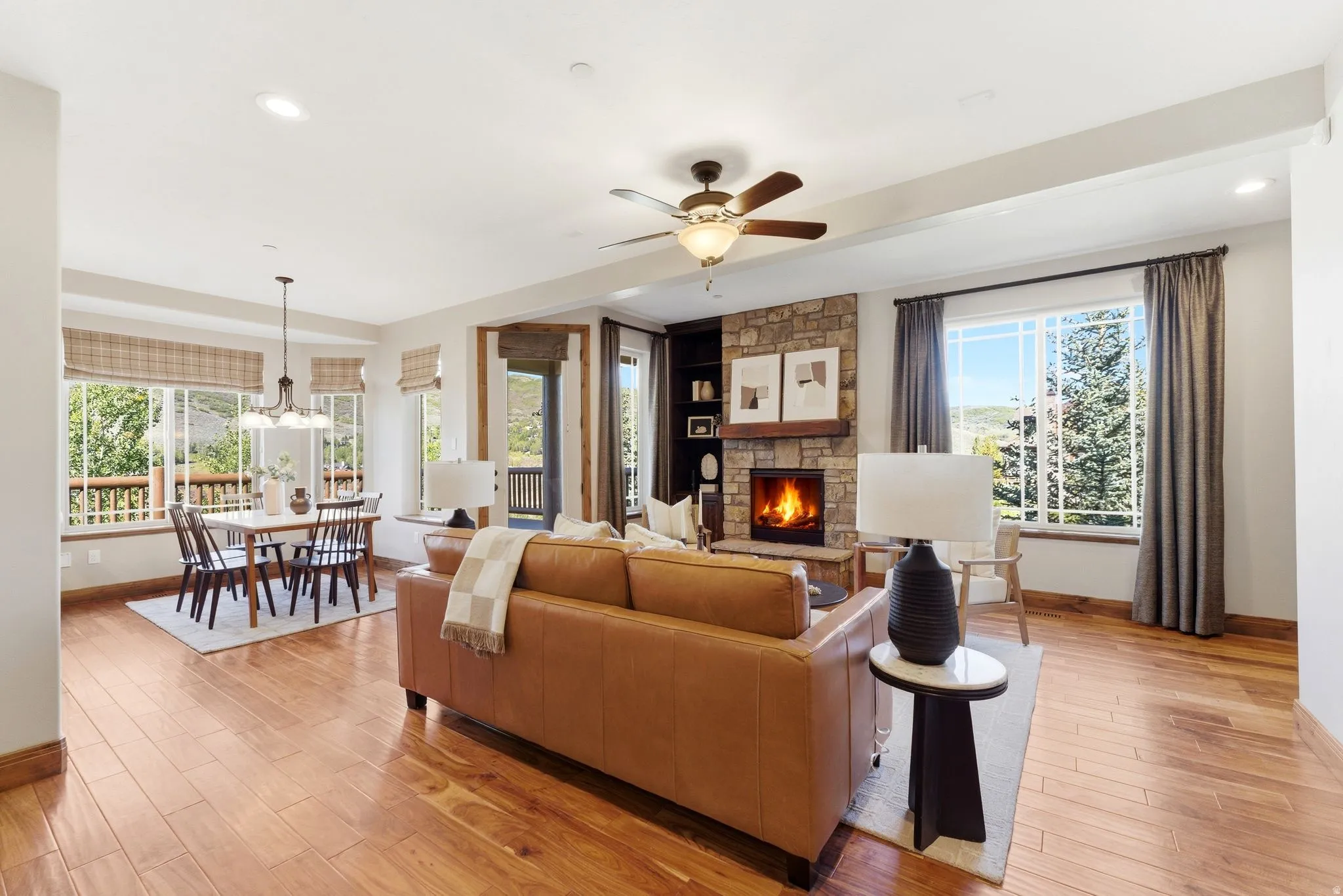 Open-concept living area featuring rich wood-finish flooring, a stone-faced fireplace with a wood mantel, a ceiling fan, recessed lighting, and multiple windows