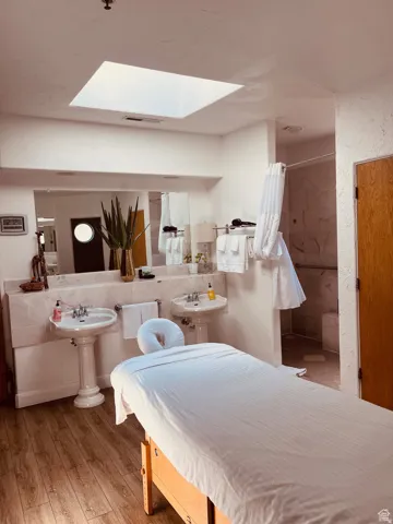 Wood-finish flooring leading to a vanity area featuring dual pedestal sinks, a large wall-mounted mirror, and a stone backsplash