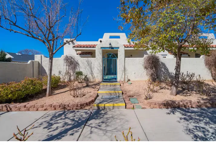 Stucco exterior with a tiled entryway, featuring a decorative iron gate and a terraced front yard with mature trees and desert landscaping