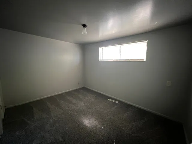 Room featuring neutral gray wall paint, dark gray patterned carpeting, a single horizontal window with blinds, and a ceiling-mounted light fixture