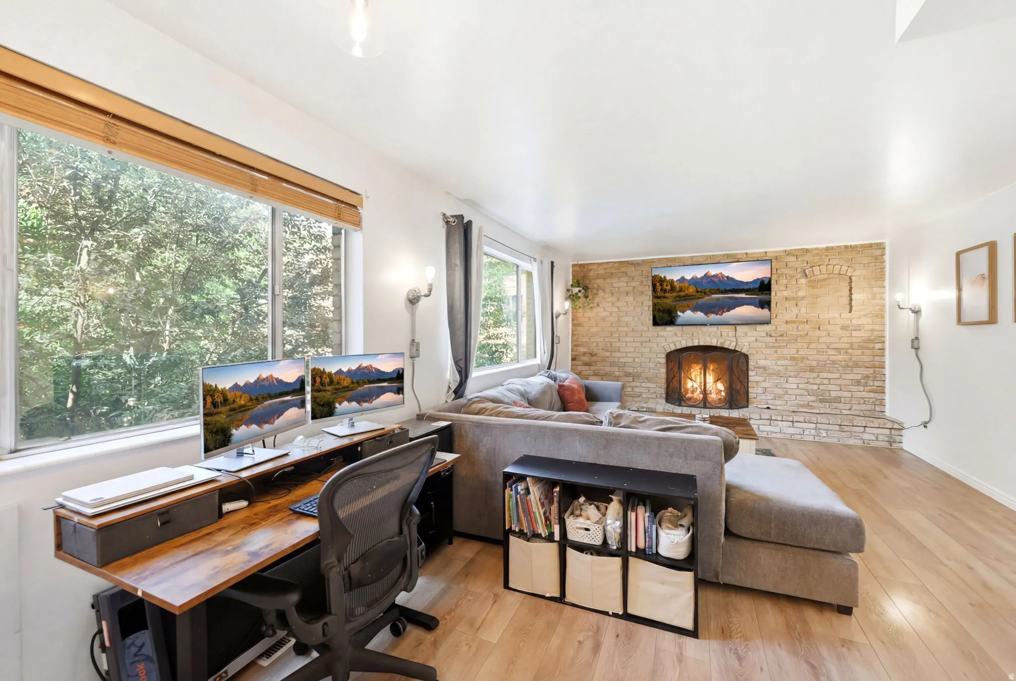 Spacious living area featuring light wood-finish flooring, a prominent brick fireplace with a hearth, and large windows