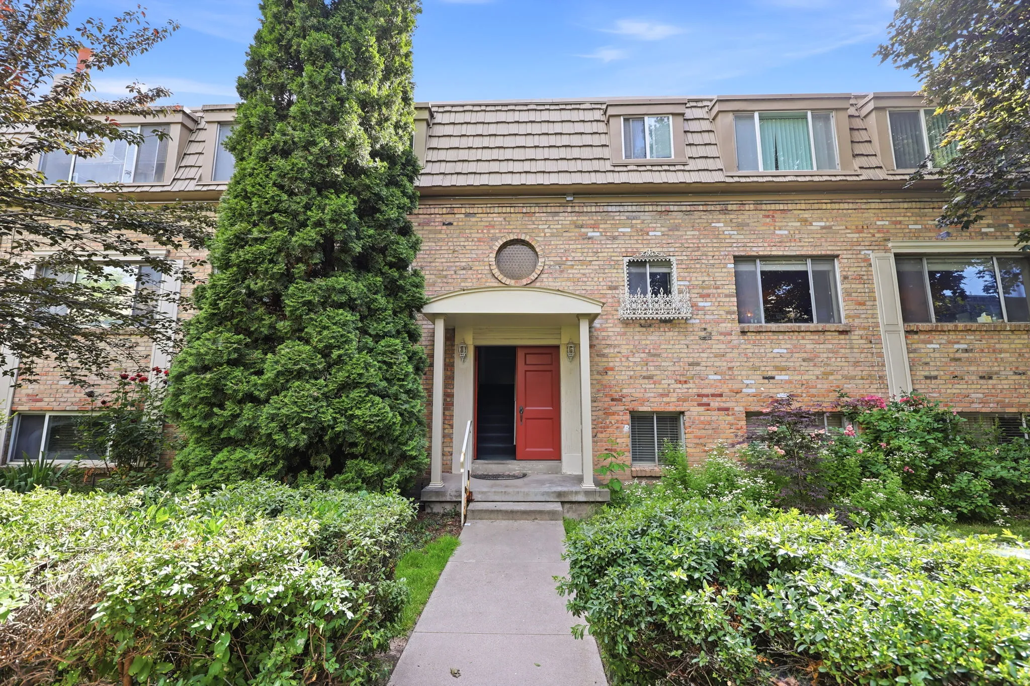Brick facade building featuring a red entry door, dormer windows, a walkway, and mature landscaping