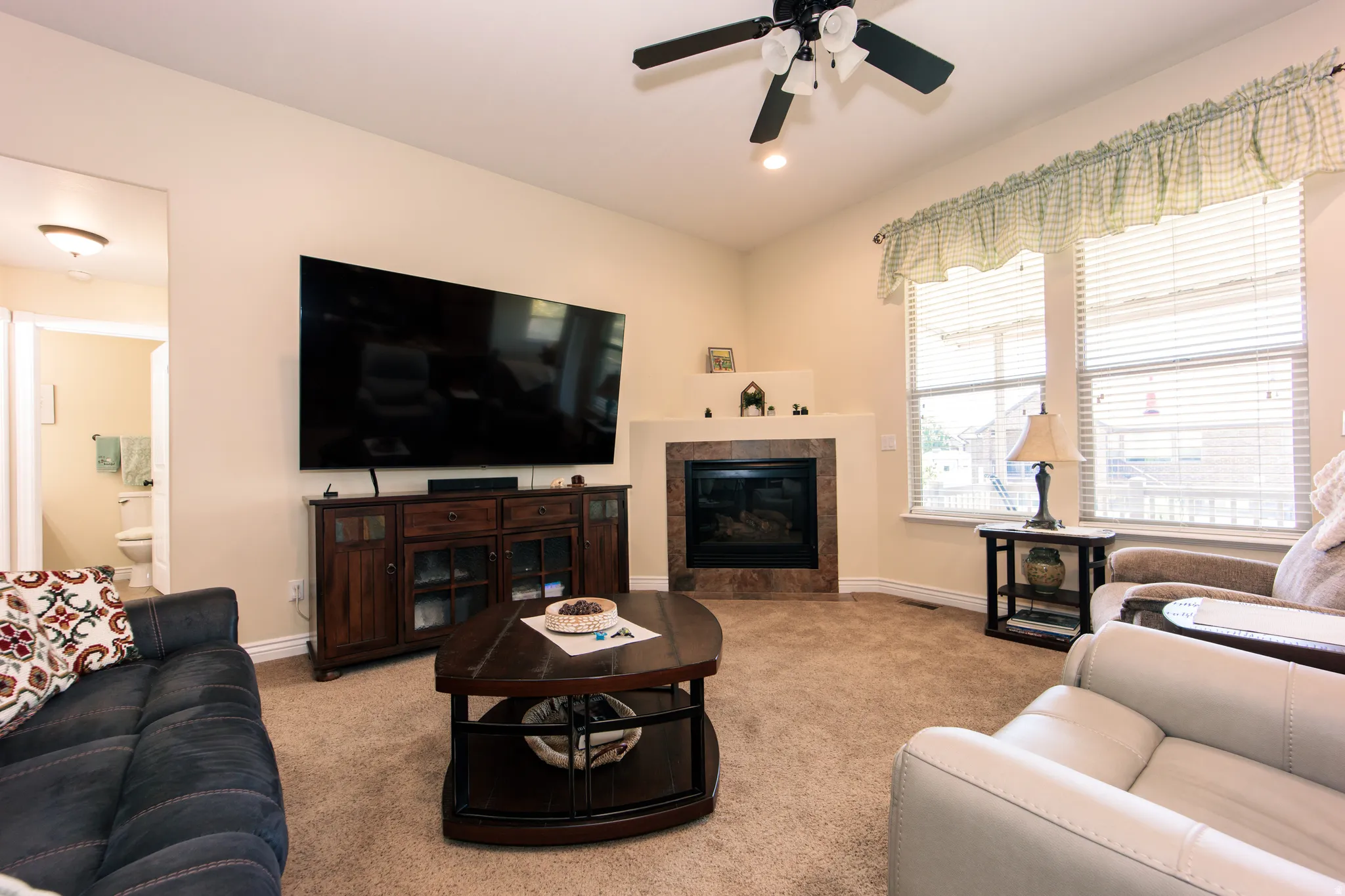 Living room featuring a corner fireplace with a dark stone surround and hearth, carpeted flooring, and a ceiling fan with integrated lighting