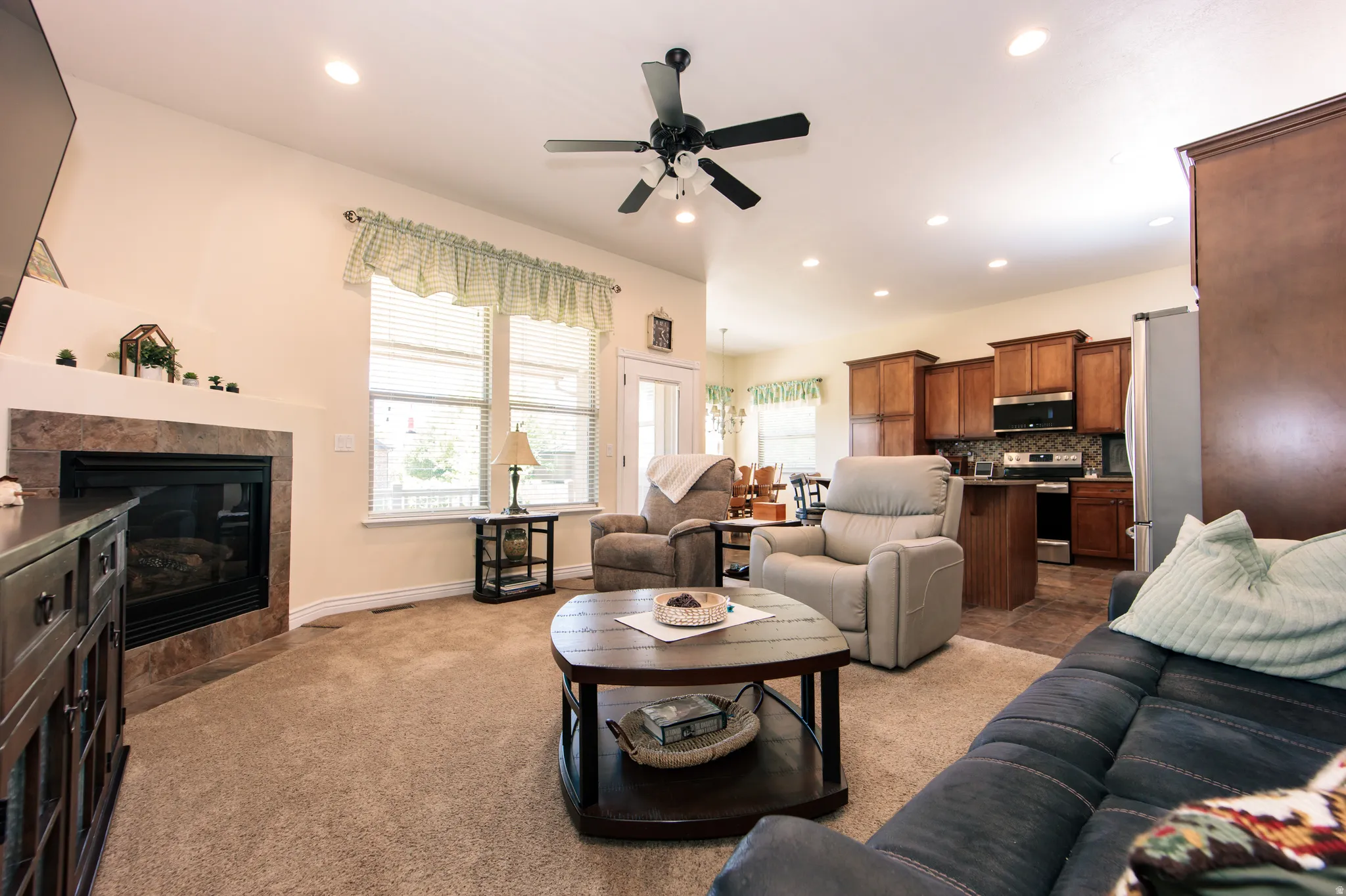 Open-concept living space featuring a gas fireplace with a stone-tile surround, neutral carpeting, and a ceiling fan with integrated lighting