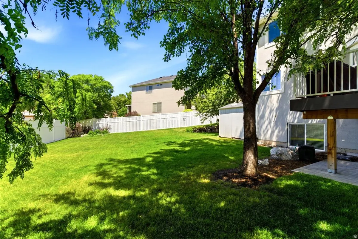 Expansive backyard featuring a lush lawn, white vinyl fencing, a mature shade tree, a ground-level patio, and a utility shed
