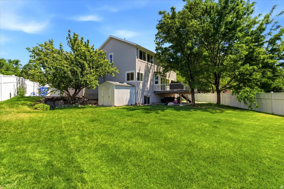 Expansive lawn area featuring a multi-level deck, mature shade trees, a white privacy fence, and an exterior storage shed