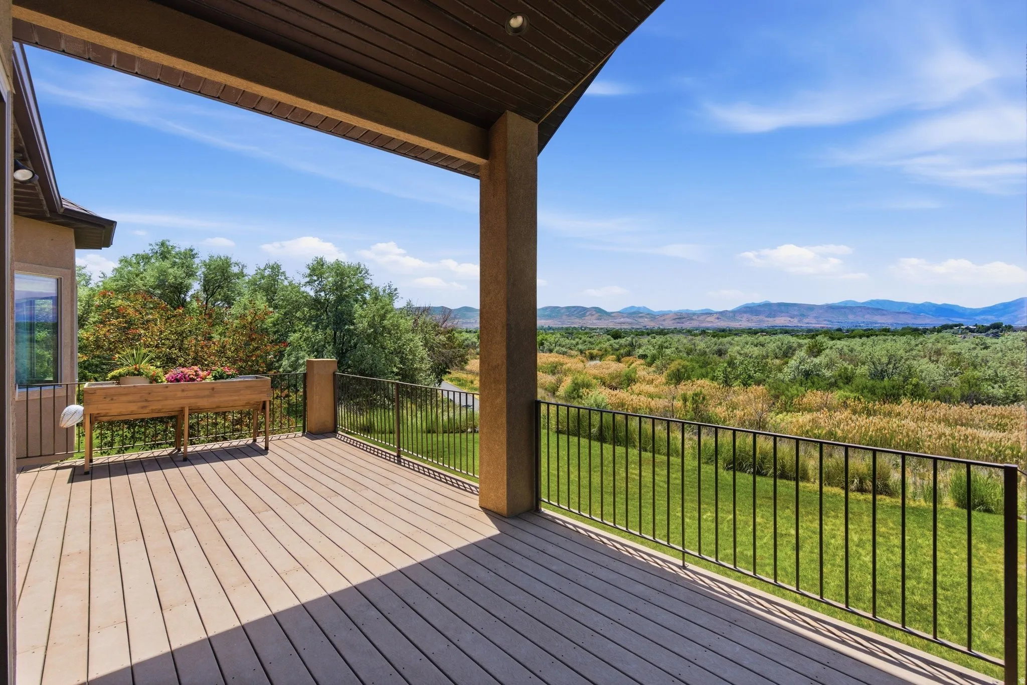 Expansive covered deck featuring composite wood-finish flooring, a stucco exterior, and black metal railings overlooking a verdant landscape with distant mountains