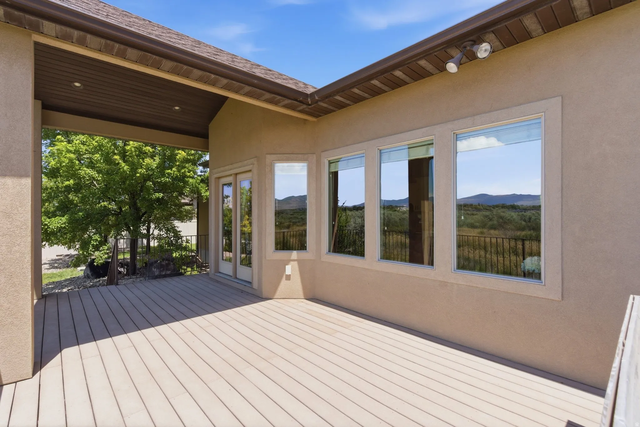Expansive outdoor deck featuring wood-finish composite decking, stucco exterior, multiple windows, double glass doors, and a covered section with recessed lighting