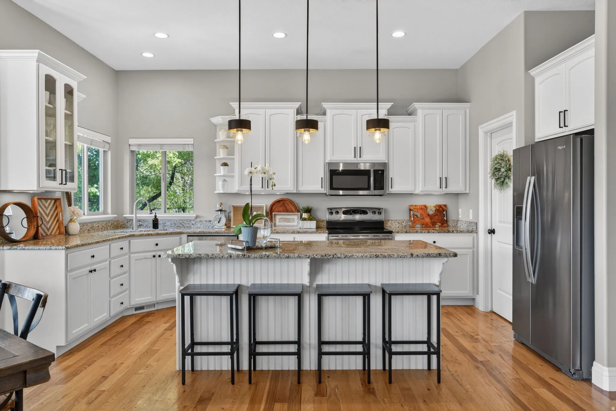 Bright kitchen featuring white cabinetry with black hardware, granite countertops, wood-finish flooring, a stainless steel microwave and range, and three pendant lights over the island