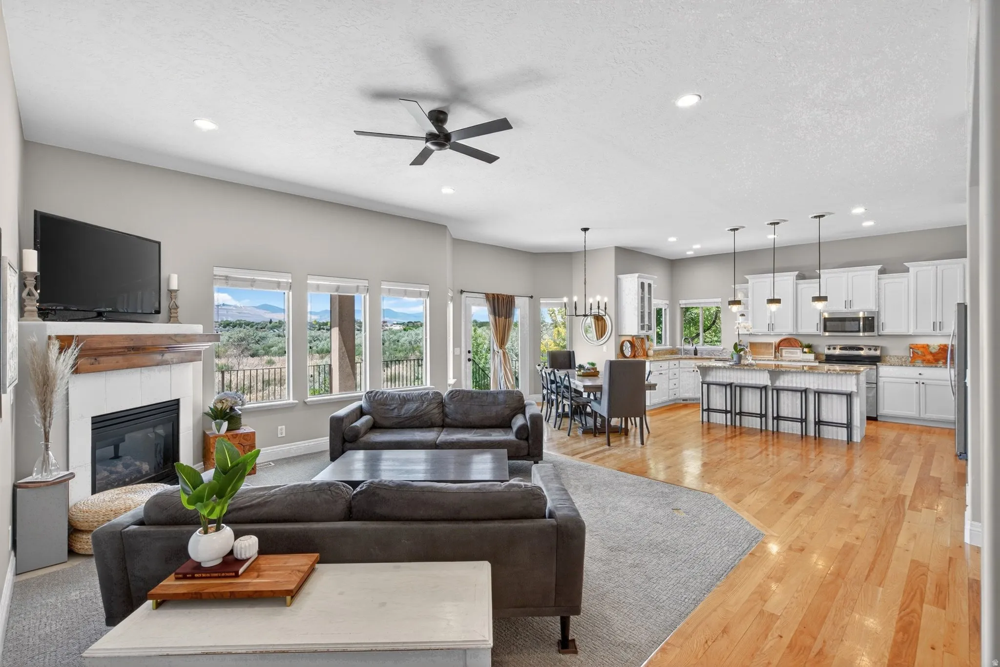 Open-concept living space featuring a fireplace with a wood mantel, multiple windows, a kitchen with white cabinetry, and wood-finish flooring