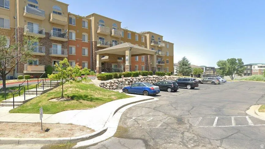 Multi-story building featuring a covered entryway, private balconies with glass railings, stone accents, and a paved parking area