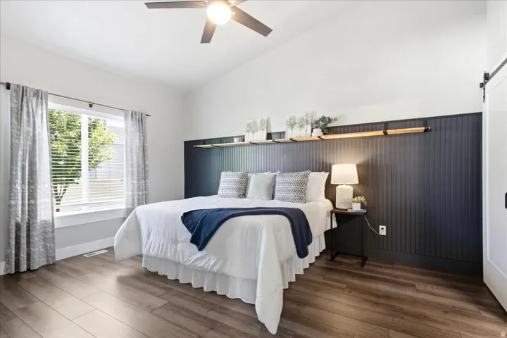 Vaulted ceiling bedroom featuring wood-finish flooring, a ceiling fan, a large window with blinds, a dark beadboard accent wall, and a sliding barn door